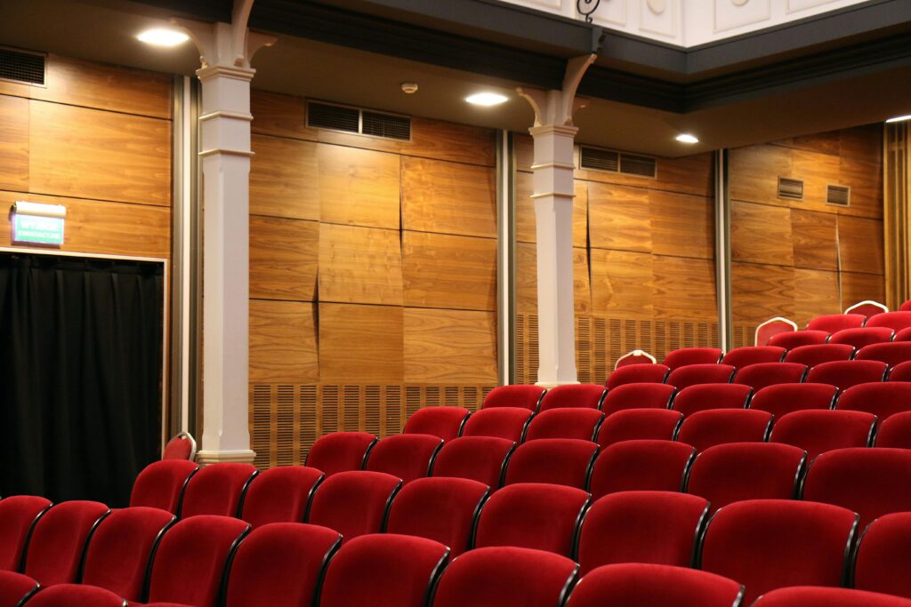 Interior of an empty auditorium with red seats and wooden panels, lit overhead.